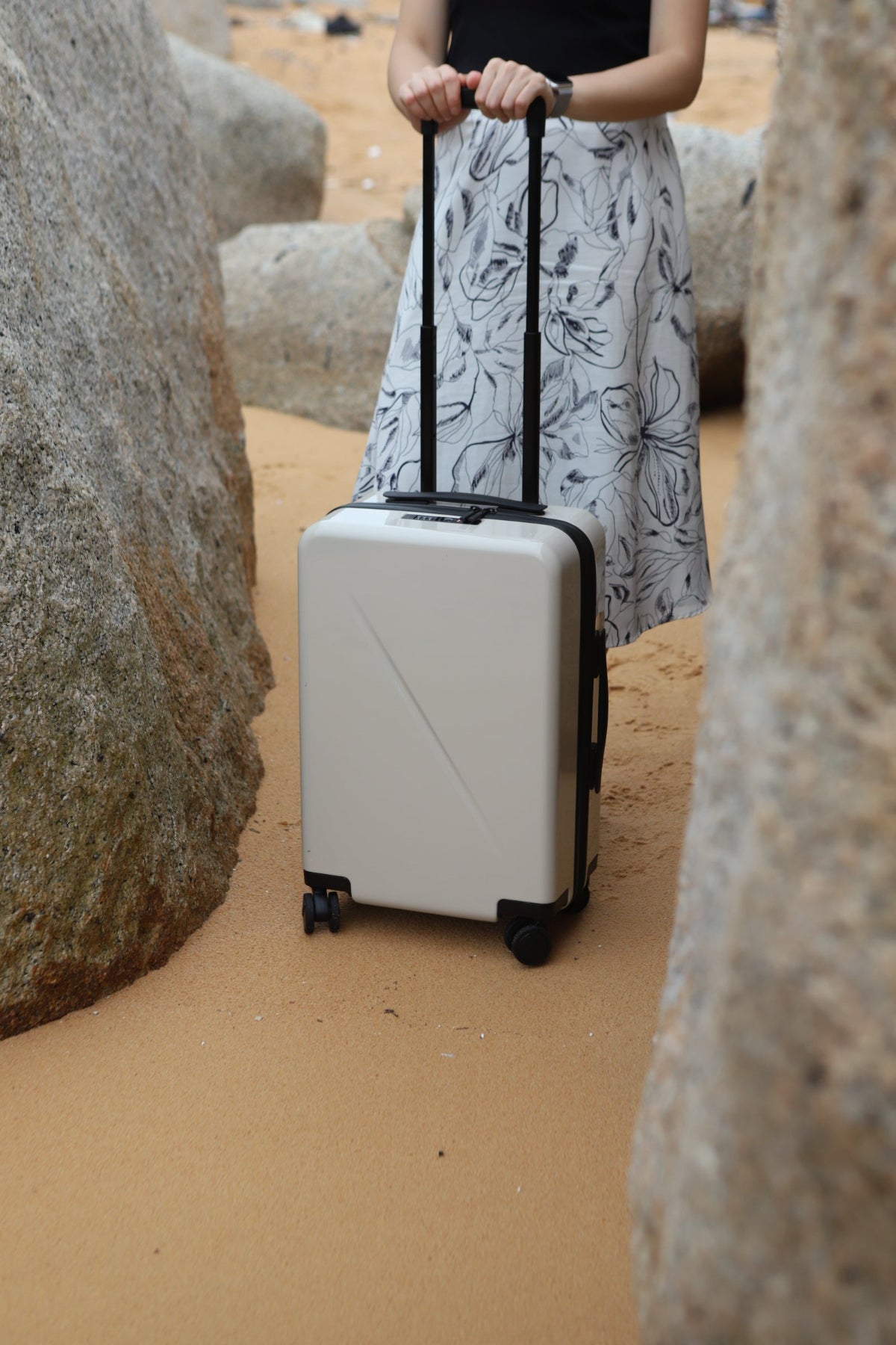 A person standing on sandy ground between large rocks, holding the telescoping handle of a hard-shell rolling suitcase. The suitcase is a  sand / off-white color with black wheels and trim.